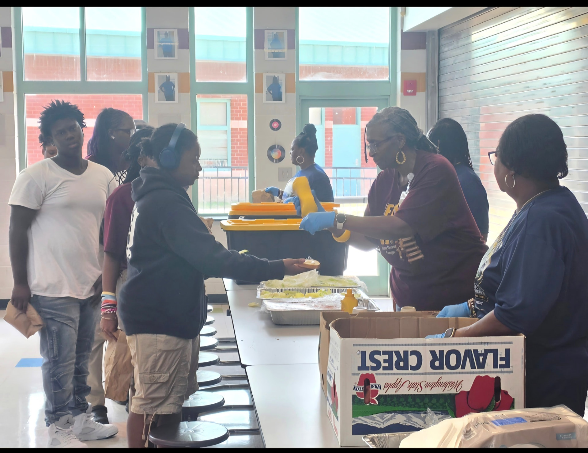 Volunteers wearing gloves hand food across a serving table to students lined up inside a bright school cafeteria.