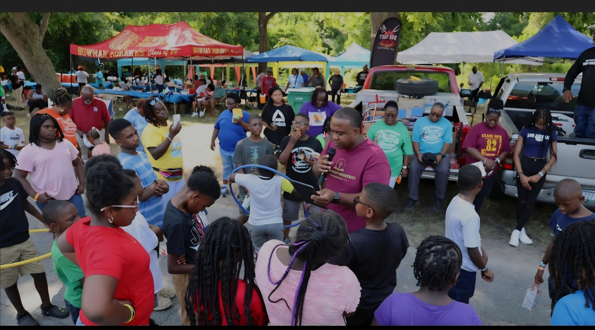 An outdoor community gathering under shade tents, where a speaker holds a microphone while children and adults stand together listening.