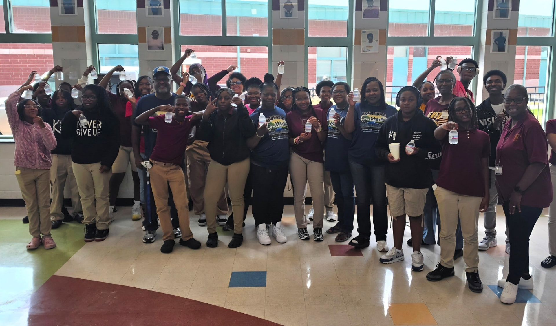 A large group of students and adults standing together indoors, smiling and raising bottled water in front of tall windows.